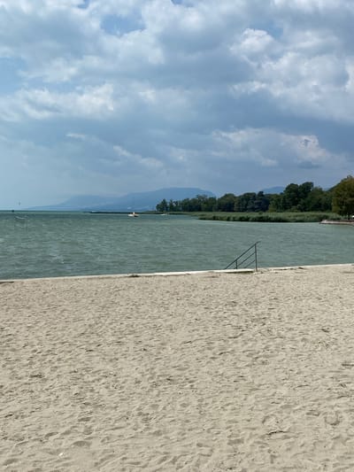 The image depicts a sandy beach leading to a body of water under a cloudy sky. In the foreground, the sand is textured with visible footprints. A short, white concrete barrier separates the beach from the water. A metal railing with several steps is partially submerged in the water. The water is choppy with visible waves. In the middle ground, reeds and trees line the shore. A small orange boat is visible on the water, along with a distant sailboat and a kiteboarder. Mountains form the backdrop. The scene is outdoors and appears to be a public beach.