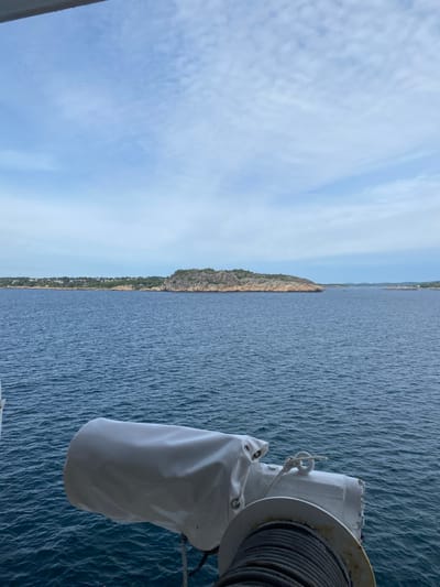 This is a vertical, eye-level, outdoor, medium shot of a coastal landscape. The image is divided roughly in half, with the left side dominated by a blue sky filled with thin, wispy clouds and the right side showing a vast expanse of blue water with gentle ripples.
In the distance, along the horizon, a rugged, rocky coastline is visible. This coastline is lined with a dense strip of green vegetation and a scattering of buildings, suggesting a settled area. The terrain rises significantly on the left side of the coastline, forming a prominent cliff face.
In the foreground, on the right side of the frame, a portion of a ship or boat is visible. A large, white, fabric-covered object, possibly a life raft or cover for equipment, hangs from a metal fixture. Adjacent to it, a large winch or spool wound with thick, dark cable is partially visible. A white rope is tied to a metal loop on the fixture.
The overall scene suggests a calm day at sea, possibly during daylight hours. The lighting indicates it could be morning or late afternoon, with soft illumination. The location appears to be coastal, with the landmass providing a visual anchor against the open water. No people or text are visible.