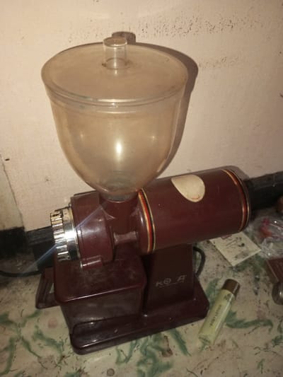 A vintage "KO A" coffee grinder sits on a tiled surface against a pink wall. It features a clear plastic hopper atop a maroon-colored grinding mechanism. A silver knob adjusts grind size. The grinder has a boxy base and a cylindrical motor housing. Beside the machine, a tube of "Body Lotion" lies on the tile, adding to the cluttered, aged aesthetic.