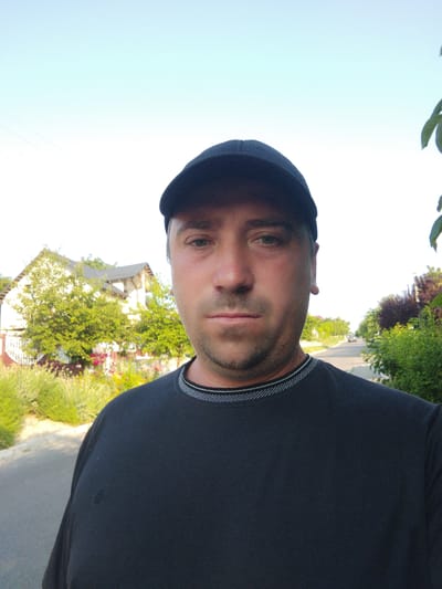 A man wearing a black t-shirt and a black baseball cap is standing outside in front of a street in the city of Costești, Moldova. He is facing the camera and has short, dark hair and stubble. Behind him, on the left side of the image, is a white house with a dark roof, surrounded by green trees and bushes. On the right side of the image, the street continues, lined with trees and more bushes. The sky is clear and bright blue.