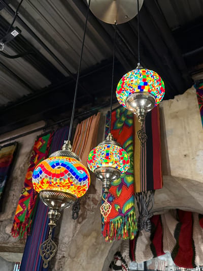 Three illuminated mosaic glass lamps hang from a circular brass-toned ceiling plate by black cords in an indoor setting within Panama City, Panama. The lamps feature distinct patterns of multicolored glass tiles. The lamp on the far left exhibits a wave-like pattern of blue, red, and orange tiles on a yellow background, while the central and rightmost lamps are spherical with a more scattered arrangement of bright multicolored tiles. All three lamps have antique brass or bronze-toned metal frames with ornate lower finials, and the central and rightmost lamps include additional decorative metal pendants. Overhead, a dark corrugated metal ceiling is visible with exposed dark metal beams and electrical conduits, including a junction box. The background features various brightly colored textiles hanging vertically, including a prominent red fabric with green and yellow floral/mandala patterns and multicolored fringe. Other visible textiles include striped fabrics in brown, orange, and black, and a lighter woven fabric with red stripes, displayed against rough, light-colored walls or architectural elements that suggest stone or stucco, possibly including an archway. No individuals are present in the scene.