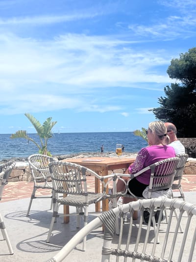 The image depicts an outdoor scene in Urbanització La Cometa, Spain, with a view of the sky, sea, and shoreline. Two people are seated in white woven chairs. A woman with blonde hair wearing a purple top leans forward, looking at something on a table. A man with white hair stands behind her. In the background, the sea is visible with the sky. Palm trees and other vegetation are present. A beer sits on the table.