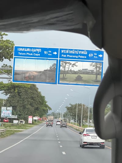 This is a rotated, vertically oriented image taken from inside a vehicle, likely a car, looking out at a road in Thailand. The primary subjects are two blue road signs, which are the main focus due to their position and clarity.

The sign on the left indicates directions to "แหลมตะลุมพุก Talum Phuk Cape" at a distance of 91 kilometers. It features an image of a beach scene. The sign on the right shows the way to "พระตำหนักปากพนัง Pak Phanang Palace" at a distance of 69 kilometers, with a picture of a building and grounds. Both signs have an upward arrow indicating the direction.

In the background, there is a multi-lane road with other vehicles visible, including a white car with a license plate showing "งบ 6489". There are trees and greenery lining the road, and streetlights are also present. Several smaller signs are visible along the roadside, some displaying numbers like "30" and "60", likely indicating speed limits or other traffic information. One sign also says "Mobil".

The scene appears to be during the daytime, with an overcast or slightly hazy sky, suggesting it might be a cloudy day or late afternoon. The overall mood is neutral, providing factual information about the location and directions. The image is partially obscured by the interior of the vehicle, particularly a dark object in the foreground which could be part of the car's interior or a hand.