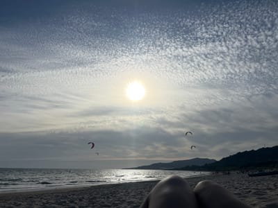 A first-person perspective shot captures a beach scene in Tarifa, Spain. The foreground shows the knees and thighs of a person lying on the sand, facing the ocean. The beach stretches out to the left, with faint figures of people and a kayak visible in the distance. The sea is calm with gentle waves, and the horizon is marked by a line of dark, rolling hills. Several kitesurfers are present, with their colorful kites dotting the sky. The sky is a blend of deep blue and white, with the sun shining brightly in the upper center, partially obscured by wispy, textured clouds.