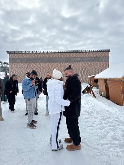 The scene is set outdoors in Yeghipatrush, Armenia, within a snow-covered environment under an overcast sky. The ground is covered in packed snow and ice. In the immediate foreground, two individuals are positioned facing each other and holding hands. The female, on the left, wears a white hooded winter jacket, white pants with black side stripes, and silver/grey winter boots. The male, on the right, wears a black fur-like winter coat, dark pants, and brown winter moccasin-style boots; he has dark hair styled in a top knot and a beard. Both individuals are smiling and looking at each other.
To the left of the central couple, a male individual is visible from the side, wearing a light blue denim jacket, grey cargo pants, and dark sneakers, holding a smartphone horizontally as if recording or photographing. Several other individuals are dispersed in the background, observing or moving within the snowy area.
In the mid-background, a large, rectangular building with patterned brick-like siding (featuring horizontal bands of brown and dark brown) stands prominently. To the left of this building, a metal structure consistent with a ski lift tower and cables is partially visible. To the right of the central couple, two small, rustic wooden cabin-like structures with snow-covered roofs are present, along with what appears to be a snowboard or similar equipment on a stand.