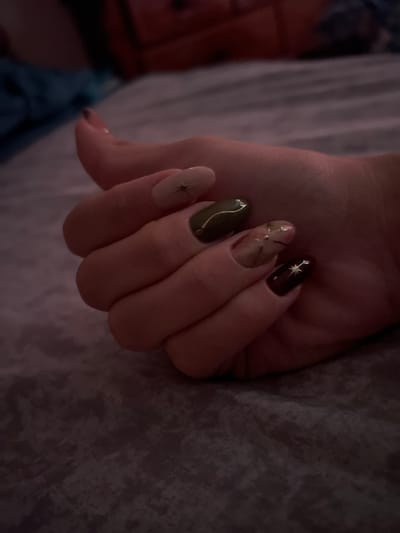 The image displays a person's hand with a manicure, resting on a textured surface. The nails are painted in various colors including a pale beige, a dark green, a lighter beige with an abstract design, and a dark burgundy. Some of the nails have a small, star-like embellishment. The background is out of focus and appears to be a bed or sofa with patterned fabric. The scene is dimly lit, suggesting an indoor setting. There is no discernible location in the city of Villa de Cura, Venezuela.