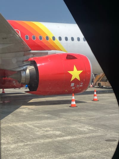 An outdoor scene on an airport tarmac in Hòa Cường Ward, Vietnam, displays the left engine, wing, and a section of the fuselage of a commercial aircraft under bright daylight. The engine nacelle is red, featuring a prominent yellow five-pointed star on its side. The engine's intake and part of its exhaust are visible. The underside of the aircraft's grey wing extends left from the engine. Above the wing and engine, the aircraft's white fuselage is adorned with horizontal stripes of red, orange, and yellow, and several passenger windows are visible. Two orange and white striped traffic cones are positioned on the concrete tarmac in front of and to the right of the engine. In the background, to the right, a yellow and brown ground support vehicle, likely an aircraft stair or loader, is partially visible, with indistinct human figures near it. The sky above is clear blue. The image is framed on the right by a dark, curved object, suggesting it was captured from an interior window.