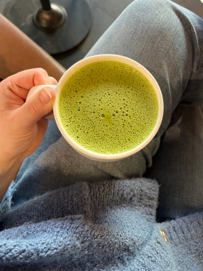 A close-up, overhead perspective shot depicts a person holding a white ceramic mug containing a bright green, frothy beverage. The froth exhibits numerous small bubbles across its surface. The person's left hand, with natural short fingernails, grasps the mug's handle. Visible below the mug are light blue denim jeans on the person's legs, partially covered by a fuzzy, blue knitted garment resting on the lap area. In the blurred background, a dark, cylindrical object, consistent with a table base or chair leg, is partially visible in the upper left, adjacent to a smooth, light brown surface. The scene is indoors. No specific identifying visual markers for the city of Kaiserslautern, Germany are present within the image.