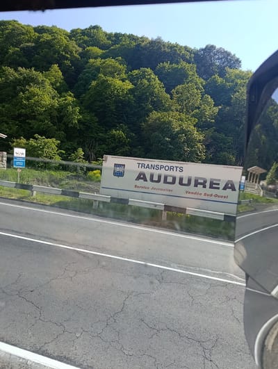The image shows a roadside scene in Semyonovka, Armenia, under a clear blue sky, suggesting daytime with fair weather. The primary focus is a white transport trailer with the words "TRANSPORTS AUDUREA" prominently displayed in large, dark lettering. Below this, smaller text reads "Service Journalier Vendée Sud-Ouest". The trailer also features a crest with stars and stripes on the left side.

To the left of the trailer, a blue sign with white text and a graphic of three horses is visible, with the words "ARMENIA'S SILK ROAD" below. The trailer and sign are positioned along a road bordered by a guardrail. In the background, a lush, dense forest covers a hillside, characteristic of a natural, possibly mountainous, environment. A small, open-air structure, resembling a gazebo or pavilion, can be seen to the right of the trailer, with a hint of another structure behind it. The road in the foreground is paved asphalt, showing some cracking, and has white lane markings. The reflection of the sky and trees is visible on the vehicle from which the photo is taken, suggesting the photo was taken from inside a car or bus. No people are directly visible in the image.
