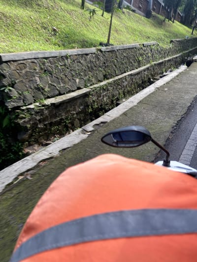 The image shows a person riding a motorcycle on a paved road. The person is wearing an orange safety vest. The motorcycle has a black rearview mirror. The road is lined by a stone wall and a ditch. The ditch is overgrown with grass and small plants. Green vegetation, likely grass, covers the hillside that rises from the road, with a few trees visible in the distance. The scene is located in the city of Kecamatan Cisarua, Indonesia, near coordinates -6.84 latitude and 107.551 longitude.