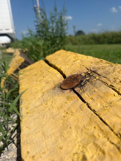 A close-up, low-angle shot focuses on a weathered, bright yellow wooden beam with a rusty metal fastener protruding from its surface. The beam is cracked and shows signs of wear and tear. Green grass grows around the beam, and gravel is visible in the foreground. In the background, out of focus, is a bright blue sky with a few scattered clouds, green foliage, and a portion of a white trailer. The overall scene suggests an outdoor, possibly industrial or rural, setting. No specific location in Cardwell, United States is discernible from the image.