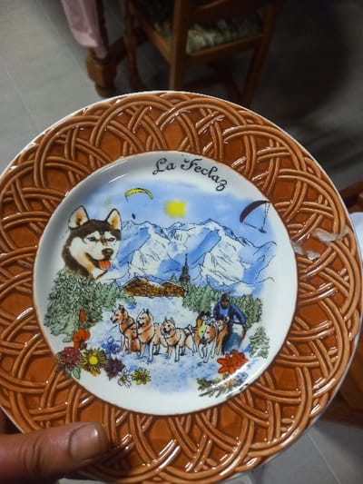 A decorative plate featuring a dog sled team scene dominates the frame. The plate's rim is woven brown. Depicted are snowy mountains, a yellow sun, a crescent moon, and a dog sled team led by a musher, set against a blue sky. A husky portrait and the inscription "La Feclaz" are visible. A human hand is on the right side of the plate. Part of a wooden chair is visible in the background.