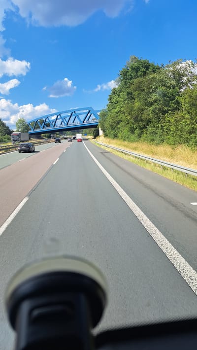 This image shows a highway scene in Haiger, Germany. The perspective is from a vehicle, with the foreground dominated by a blurry, dark object, possibly a dashboard-mounted device. The highway itself stretches into the distance, with lanes marked by white lines. Several vehicles, including a truck and cars, are visible on the road. A prominent blue truss bridge spans across the highway in the mid-ground. To the right of the highway, a grassy embankment with a metal guardrail is visible, leading into dense green trees. The sky is a bright blue with scattered white clouds.