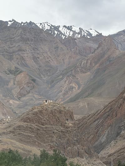 The image shows a wide, mountainous landscape under a cloudy sky. The mountains are rugged and appear to be barren with patches of snow on the higher peaks. In the mid-ground, perched on a rocky outcrop, is a small cluster of white buildings that resemble a monastery or a settlement. This settlement is likely the Zangla monastery, located in the Zangla village of Zanskar valley, Ladakh, India. The lower part of the image shows some green vegetation, likely trees, suggesting a lower elevation in the immediate foreground. The overall impression is one of a remote, high-altitude, and possibly ancient settlement nestled within a dramatic mountain range.