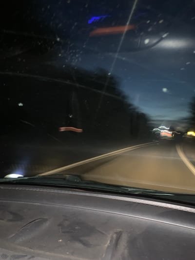 The image depicts a view from inside a vehicle, looking forward through the windshield at a rural road at night. The road is illuminated by headlights, and trees line either side. The sky is dark, with a few faint lights visible. The interior of the car, including the dashboard, is partially visible in the foreground. There are no people or animals visible in the scene. The overall impression is one of driving on a quiet road in a remote area. There is no information in the image that allows for the identification of the specific location as Zolotovo (Sitnikovskii S/S), Russia.