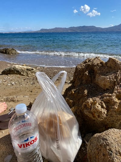 The image depicts a static scene on a rocky and sandy beach, specified as Megalochori, Greece. In the foreground, a clear plastic bottle of "VIKOS Natural Mineral Water" with a white cap stands upright next to a translucent plastic shopping bag containing light-colored objects. Both items rest on the sand, adjacent to a large, textured brown rock formation on the right. A bare human left foot is partially visible in the lower-left quadrant. The immediate background consists of calm, blue-green sea water with small waves breaking softly on the shore. Across the water, a distant landmass characterized by hills or mountains is visible under a clear blue sky with a few scattered white cumulus clouds.