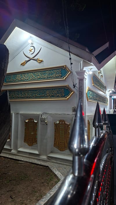 A low-angle, medium shot captures the exterior of a white building at night, likely a religious site, adorned with Islamic calligraphy and symbolism. Two crossed scimitars, a star, and a crescent moon are mounted on the gable end of the building. Three horizontal panels with gold Arabic calligraphy on a teal background are affixed to the white wall. Below these, two large gold decorative panels with intricate patterns are set into the wall. The building's facade is illuminated by artificial lighting, casting shadows. In the foreground, a blurred silver railing with pointed finials suggests a barrier or entrance. The ground in front of the building is covered with dirt and some sparse vegetation, bordered by a patterned tiled pathway. The overall scene is dark, with the exception of the illuminated building and a few scattered lights. This location is identified as Lhokdagang, Indonesia.