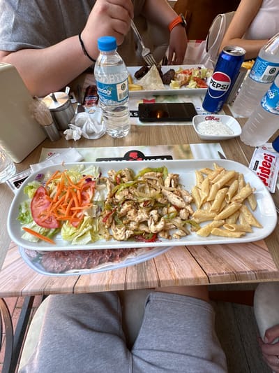 An overhead view of a dining table in a casual restaurant setting in Edirne Merkez, Turkey, is depicted during daytime. Three individuals are partially visible. On the left, a person wearing a light gray short-sleeved t-shirt shows their left forearm with a black string bracelet and their hand holding a fork over a white plate. On the right-back, another person, wearing a white top, shows their arm and hand with an orange wristwatch, also holding a fork. The person at the bottom of the frame has their legs visible, clad in light grey sweatpants, with a light-colored shoe on one foot.

The central focus is a large, white rectangular plate containing three distinct food items: a salad of shredded lettuce, sliced tomatoes, and julienned carrots; sautéed chicken strips with green peppers and onions; and penne pasta in a light-colored sauce. A second white plate, positioned further back, holds meatballs, a piece of flatbread, and French fries.

Beverages on the table include a blue Pepsi can, a 0.5-liter "Buzdağı" brand bottled water, and two "Karya" brand 500ml bottled waters. Additional table items consist of a small white dish of "ÇITIR USTA" branded white dipping sauce, a stainless steel shaker, a container with plastic-wrapped toothpicks, a black smartphone lying face down, a red packet of "ÇITIR USTA" branded sauce, and crumpled paper napkins. The table is wood-textured, covered with placemats displaying a wood grain pattern and Turkish text, including a disclaimer about visuals being for advertising purposes. The floor beneath the table is dark.