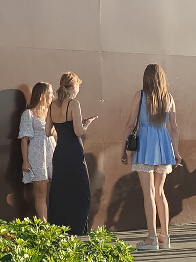 Three young women are standing outside in front of a brown wall, likely in an urban or suburban setting. The woman on the left is wearing a white, short-sleeved dress with a floral pattern. She is holding a white object, possibly a phone or a small bag, in her left hand. The middle woman is wearing a long, black dress with spaghetti straps. She is looking at her phone, which she is holding in her right hand. The woman on the right is wearing a short, light blue dress with white lace trim at the hem. She is holding a lit cigarette in her right hand and has a black handbag slung over her left shoulder. The women appear to be engaged in conversation or looking at their phones. The environment suggests a warm day, with sunlight casting shadows on the wall. Green foliage is visible in the foreground. The specific location in Sint-Pieters-Leeuw, Belgium, is not discernible from the image itself.