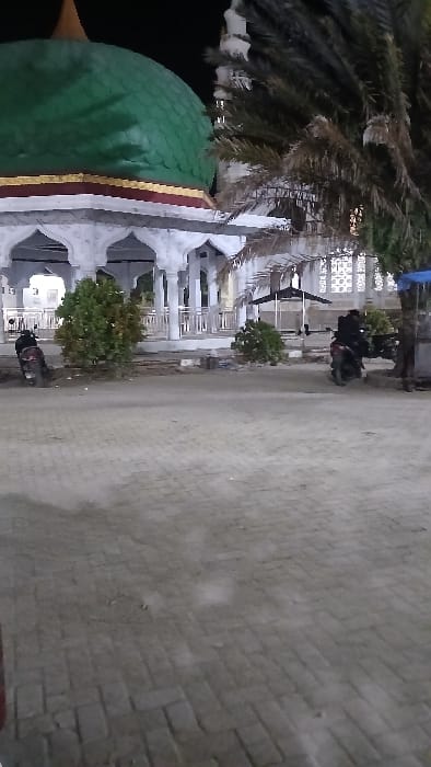 The image depicts an exterior view at night in RawangItiek, Indonesia, focusing on a building with Islamic architectural elements. The dominant subject is a structure featuring a large, smooth green dome with a gold and maroon band at its base. This building, likely a mosque, has white or light-colored pillars forming an arcaded portico beneath the dome. Behind and to the right of the dome, a tall, slender white minaret is partially visible against the dark sky. Further to the right, behind a palm tree, the building displays white lattice-work or decorative window screens.

In the mid-ground, a paved area constructed from interlocking bricks or pavers extends across the frame. Two dark-colored motorcycles are parked on this paved surface near the base of the main building. One motorcycle is positioned on the left, near a small green bush. The second motorcycle is located further right, adjacent to another small bush and near a partially visible blue tarp or awning. A black, open-sided tent-like structure is situated further back on the right side of the scene. Several palm trees are present, with one prominent tree dominating the right mid-ground, its fronds partially obscuring parts of the background. The scene is illuminated by artificial light sources, creating moderate visibility against the dark night sky. No individuals are discernible in the frame.