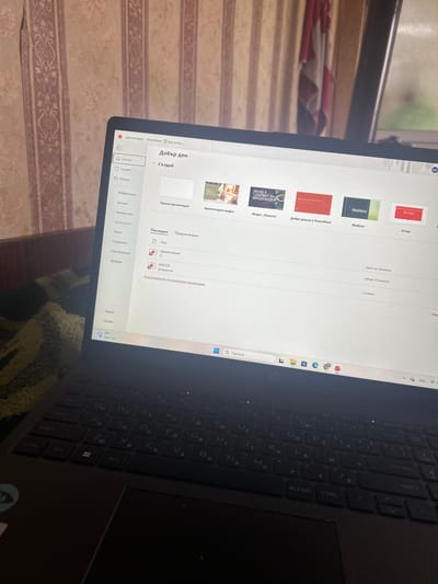 A close-up shot shows a laptop screen displaying Microsoft PowerPoint in Bulgarian. The open PowerPoint page titled "Dobur den" (Good day) lists recent documents like "Atlas", "Madison", and "Добре дошли в PowerPoint" (Welcome to PowerPoint). The interface includes options like "Създай" (Create), "Отвори" (Open), and file management options. The laptop's keyboard is partially visible. The background shows a patterned surface, likely a couch or tablecloth, and a hint of an exterior scene through a window. The image's precise geographic location cannot be determined based on the provided information.