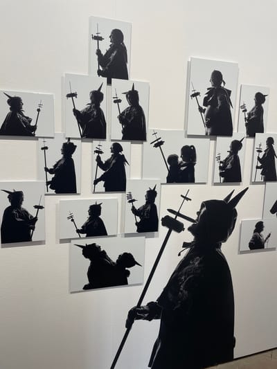 The image depicts an art installation consisting of numerous black and white silhouette photographs displayed on a white wall, suggesting an exhibition space. The photographs feature various figures, primarily women and children, in profile, holding staffs topped with decorative elements. A large, prominent silhouette of a figure wearing a horned hood and holding a staff is positioned in the foreground, partially obscuring some of the smaller photographs. The background is a plain white wall, consistent with a gallery setting. This installation is located in the city of Mataró, Spain, specifically within the Museum of Mataró.
