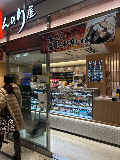 A storefront of a shop, likely a bakery, located in Chiyoda City, Tokyo, Japan. The shop features a glass display case filled with pastries and goods. A person wearing a brown jacket and black pants walks past the store, with their back turned towards the camera. The person is walking from right to left in the scene. The storefront also has a large sign with Japanese writing on it, hanging over the entryway. The storefront has a wooden frame and a tiled floor. The store is surrounded by other buildings and appears to be located in a busy commercial district. The store's signage is in Japanese, and the sign is on the left side of the entrance, hanging vertically. The storefront has a large glass door with a glass panel above it. The glass panel is also adorned with Japanese writing. The glass door opens to a tiled floor. The storefront is located on a street corner with a busy street in the background. The street is filled with pedestrians and vehicles. The building is made of concrete and glass. The signage is in Japanese, and it is not possible to translate it without further context.