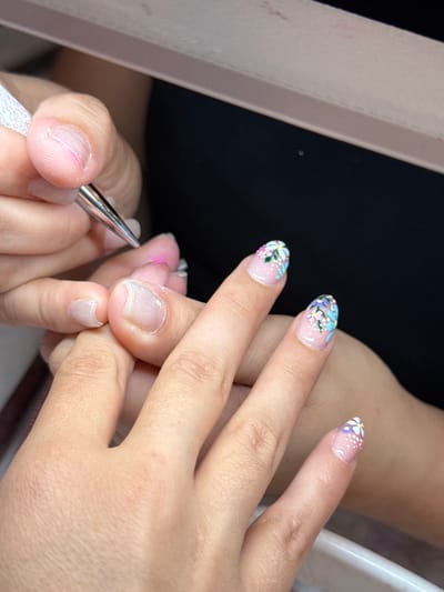 The image depicts a close-up, high-angle view of two individuals' hands engaged in a nail art application, consistent with a professional salon setting in Naples, United States. In the foreground on the left, a hand with fair skin, belonging to an unseen individual, holds a fine-tipped metallic nail art brush with a textured white handle. This hand is positioned to apply a translucent pink substance onto the pinky fingernail of a second individual's hand, which occupies the center and right side of the frame.
The second individual's hand, also with fair skin, displays multiple fingernails in various stages of a manicure. The pinky fingernail has a clear gel base, and a thin line of pink is being meticulously drawn across its surface by the brush. The ring finger, middle finger, index finger, and thumb of this hand are visible. The middle and index fingernails feature elaborate floral designs with pastel colors including light blue, purple, pink, yellow, and white, applied over a clear or light pink gel base, accentuated by small white dots. The thumbnail also exhibits a completed floral design.
The background is dark and out of focus. A light-colored, possibly white or light grey, flat overhead panel or light fixture is partially visible at the top of the image, providing direct illumination to the hands and work area. The interaction involves a precise artistic application of nail embellishment.