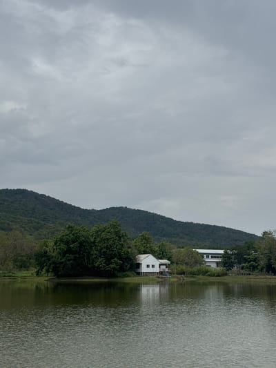 The photograph shows a lakeside view with buildings near the water's edge, set against a backdrop of a forested hillside and an overcast sky. The water is calm, with a visible ripple pattern across its surface. There are two visible buildings, both white, positioned near the lakeside and among trees. The hillside is densely covered with green vegetation, and the sky is filled with grey clouds. The photo appears to be taken near Ban Huai Sai, Thailand.