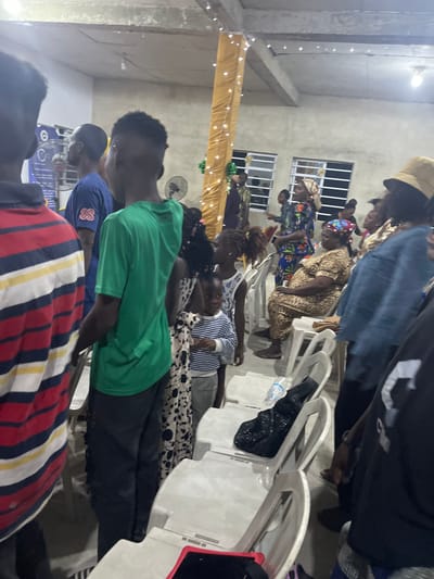 A group of people are seated in rows of white chairs, likely in a church or meeting hall. Some are wearing colorful clothing and head coverings. Decorations adorn the room including a gold curtain and string lights. A ceiling fan is also visible. People's heads are bowed.