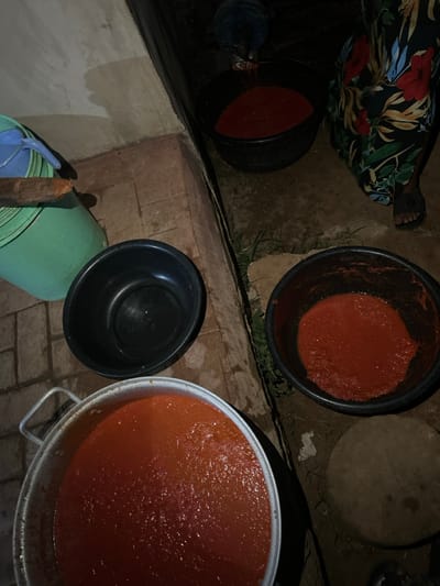 Four buckets and a large pot filled with red sauce are arranged on a brick surface, outside near a grassy area. One bucket is empty, sitting atop a green plastic container. The sauce appears consistent in each container. A person's legs and patterned clothing are visible in the top left corner. The setting is dimly lit.