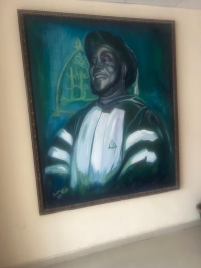 The image shows a framed painting hanging on a white wall. The painting is a portrait of a Black man with a beard, wearing a hat and a white shirt with dark stripes. There appears to be a green emblem on the shirt. Behind the man, there is a faint depiction of a musical instrument, possibly a saxophone or trumpet. The background of the painting is a blend of blue and green hues. The frame is dark brown and ornately decorated. The signature of the artist, Amima X One, and the year, 2014, can be seen in the bottom right corner of the painting. The location is specified as Jama A, Nigeria.