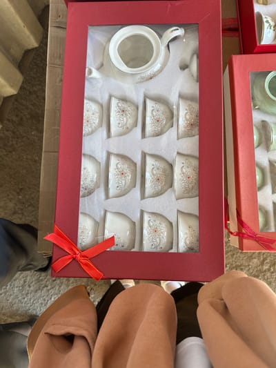 The image shows a gift box containing a set of white porcelain teacups and a teapot, decorated with intricate silver and red floral patterns. The box has a red exterior and a clear plastic window. A red ribbon is tied in a bow on the front of the box. To the right of the main box, another gift box, also red with a clear window, is partially visible, containing more teacups. The boxes are placed on a textured surface, possibly a carpet or a mat. The background shows glimpses of a wooden surface and some fabric. The overall scene suggests a retail or domestic setting where the tea set is being presented or stored. The image is taken from a high angle, looking down at the boxes and a portion of what appears to be people's legs and feet clad in socks and leggings. The location is specified as Kara-Suu, Kyrgyzstan.