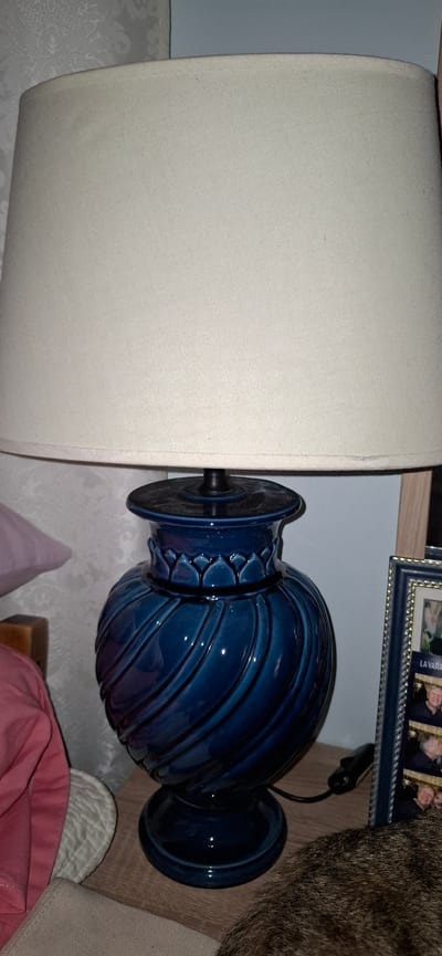 This is a close-up, eye-level shot of a decorative blue ceramic lamp with a textured, cream-colored lampshade, placed on a wooden surface. The lamp has a swirling, ribbed design and a fluted decorative element just below the shade. To the right of the lamp, a framed picture is visible, containing a strip of four photographs of two individuals, with some text partially visible at the bottom. The background shows a textured wallpaper. The setting appears to be an indoor environment, possibly a bedside table or a side table in a room in Aubagne, France.
