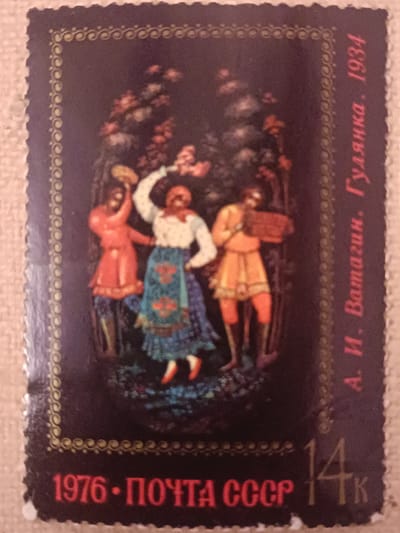 The image is a close-up, slightly blurred photograph of a postage stamp. The stamp depicts a folk art scene with three figures in traditional attire dancing in a forest setting. The figures are a man in an orange tunic and hat, a woman in a white blouse, a patterned blue skirt, and a red headscarf, and another man in a red tunic and dark pants. The background features trees with red and pink blossoms. The stamp has a decorative border. Text in Russian is visible on the right side of the stamp, reading "А. И. Ватагин. Гулянка. 1934". Along the bottom, the text "1976 • ПОЧТА СССР" and "14к" are present. There is no indication of Onaldy, Turkmenistan in the image or any context provided.