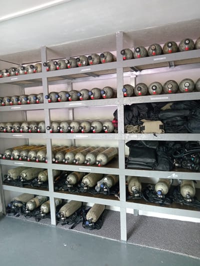 In a storage room, likely in Aldan, Russia, several shelves are filled with scuba diving equipment. The shelves, made of metal, are packed with rows of compressed air tanks. These tanks are predominantly silver or grey, with some featuring yellow markings and others covered in fabric bags. Each tank has a regulator attached, with a red or blue cap on the valve. In some of the lower shelves, black fabric bags are neatly stacked, possibly containing diving gear. The room itself appears utilitarian, with plain walls and a light-colored ceiling. The floor is grey concrete, with a patterned grey matting present on some of the lower shelves. No individuals are visible in the image.
