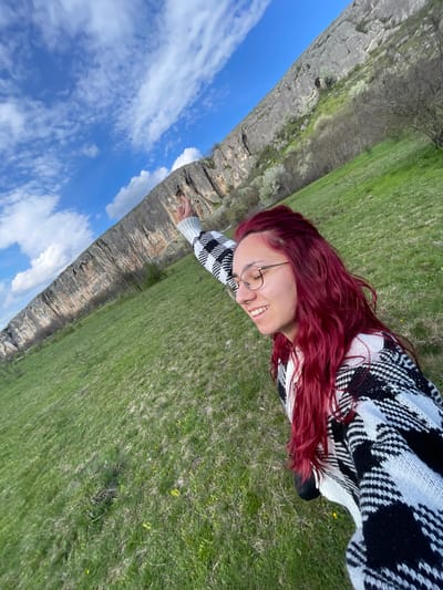 A woman with red hair and glasses is lying on a green grassy field next to a rocky cliff face. The sky is visible in the background with white clouds. The woman is wearing a black and white sweater and appears to be smiling. The image appears to be taken from a slightly overhead angle and is rotated counterclockwise. The location is Barkach, Bulgaria, which is likely the location of the cliff formation.