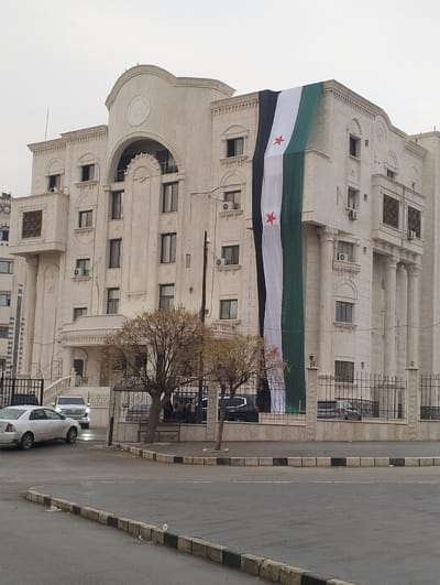 A large cream-colored building with a prominent Syrian flag draped vertically down its right side dominates the scene. The building has multiple floors, windows, and architectural details, including columns and an arched entrance. In the foreground, a curved street with black and white edging is visible. Several cars are parked or driving on the street, partially obscured by leafless trees. A fence surrounds the building, creating a barrier between it and the street. The sky is overcast.