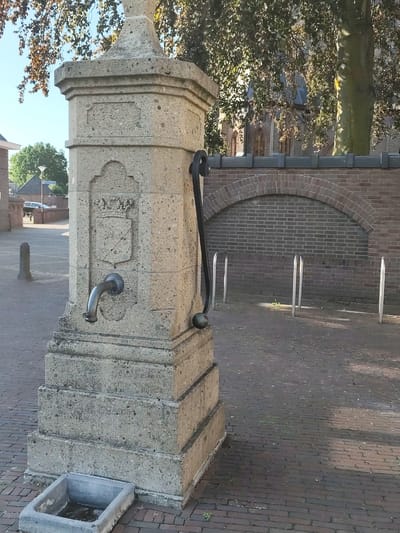A stone water pump stands in a paved area in front of a brick wall with an arched recess. The pump is made of concrete blocks and has a metal spout and handle. A small concrete basin is located at the base of the pump. In the background, trees with autumnal foliage are visible, along with a building with a steep roof. The scene is outdoors on a sunny day, likely in the morning or afternoon, given the shadows. The location is Schaijk, Netherlands. There is no visible text in the image.