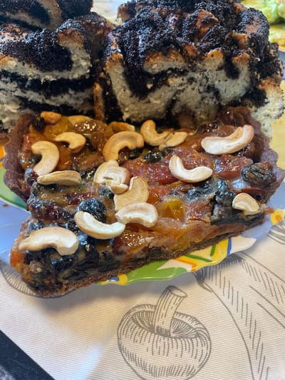 A close-up shot shows a slice of fruit tart with cashews, sitting in front of a poppy seed cake. The tart has a dark crust and is filled with an assortment of dried fruits, including raisins and possibly apricots, all coated in a glossy glaze. Several cashews are artfully placed on top. The poppy seed cake is visible in the background, with distinct layers of bread and poppy seed filling. The food is presented on a white tablecloth with a subtle pattern of apples. The setting appears to be indoors, likely a domestic kitchen or dining area. There is no indication of the specific location within the city of Турово, Russia.