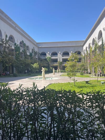 A bright, symmetrical courtyard is flanked by light-colored buildings with arched balconies. Lush greenery covers the ground and some building facades. A pathway stretches through the courtyard, dotted with statues and trees. The sky is clear blue, with a large bush obscuring part of the view.