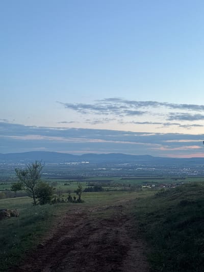 The scene presents a landscape viewed from a high vantage point in Balsha, Bulgaria, near the vicinity of the Balkan Mountains.
**Sky:** The left portion of the image shows a light blue sky, transitioning to streaks of grey, white, and pink clouds.
**Mountains and City:** A distant range of mountains forms a barrier above the city. The cityscape below is a mixture of residential and commercial buildings.
**Vegetation:** The right side of the image depicts grassy fields and trees. A brown dirt pathway runs through the field.