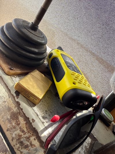 The image displays a collection of objects placed on a textured surface, possibly a table or a boat deck, given the Mikołajki, Poland location context. The dominant subjects are a bright yellow and black walkie-talkie, a brass Zippo lighter, a black weight plate with a threaded metal rod attached, and a pair of dark-rimmed sunglasses with a red accent visible. The walkie-talkie has a grid of ventilation holes and buttons, with the brand name "Motorola" partially visible. The Zippo lighter has text inscribed on it, including "BRADFORD, PA. MADE IN U.S.A.", and "ZIPPO". The weight plate appears to be made of black rubber or plastic and is stacked in several layers.
The lighting suggests it's daytime, with sunlight casting shadows and highlighting the textures of the objects and the surface. The surface itself has a weathered or distressed appearance, with peeling paint or material in some areas, revealing brown undertones. The scene is static, with no activity depicted. The overall impression is one of personal items laid out, perhaps before or after an outdoor activity. The objects are arranged closely together, creating a still-life composition.