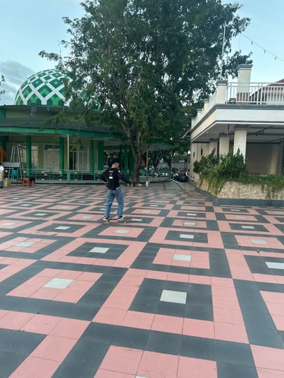 In Bantaeng, Indonesia, on a sunny day, a young man in a black long-sleeved shirt and blue jeans is skateboarding on a patterned courtyard. The courtyard features a repeating design of pink and dark grey tiles, with lighter grey squares interspersed. To the left, a green building with white window frames and a distinctive green dome with a white lattice pattern, likely a mosque, is visible. A large tree with dark green foliage stands between the courtyard and the mosque. To the right, a white building with columns and a railing on an upper level is partially visible. The sky is a pale blue with some clouds. In the background, several motorcycles are parked, and string lights are suspended overhead.