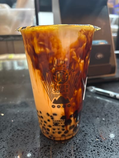A clear plastic cup containing a brown sugar boba tea from "Tiger Sugar" is centered on a dark, speckled countertop. The beverage features vertical streaks of dark brown sugar syrup against a lighter, opaque beige liquid, with black tapioca pearls settled at the bottom. The cup is sealed with a clear plastic film and bears the "Tiger Sugar" logo, which includes a tiger's face icon, the text "BRAVE AS A TIGER," and Chinese characters. In the blurred background, behind the cup, various indistinct objects and surfaces suggest an indoor commercial setting, possibly a bubble tea shop in Winchester, United States of America. No individuals are visible.