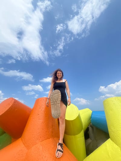 A young woman with curly brown hair is standing on brightly colored, concrete tetrapods against a blue sky with white clouds. She is wearing a black slip dress and dark sandals, and her right foot is raised, showing the sole of her shoe. The tetrapods are predominantly orange, yellow, and blue, arranged in a cluster that appears to form a breakwater or barrier. The perspective is from a low angle, looking up at the woman and the sky. The scene suggests an outdoor environment, likely a coastline or harbor area. It is not possible to determine the specific location within Phú Thủy, Vietnam based solely on this image.