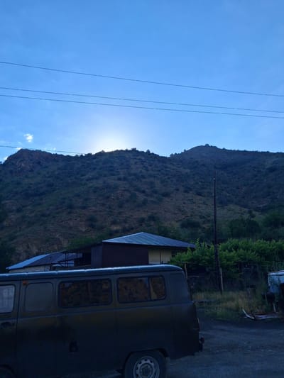 The image depicts an outdoor scene under a clear blue sky with the sun partially obscured by a distant mountain range. In the foreground, a dark-colored van is parked on a dirt road. Behind the van, several buildings with corrugated roofs are visible, nestled at the base of a vegetated mountain. Power lines stretch across the upper portion of the frame. The overall setting suggests a rural or semi-rural environment, likely in Vayk, Armenia, given the mountainous terrain and architecture. There are no people or discernible actions taking place in the image.