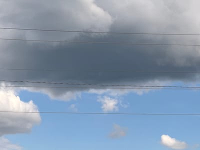 A wide-angle shot captures a segment of the sky, divided by three parallel utility wires stretching horizontally across the frame. The sky itself presents a dramatic contrast: a heavy, dark gray cloud mass occupies the upper portion, casting a somber mood, while the lower portion reveals a bright blue sky dotted with fluffy white clouds. The wires, appearing as thin, dark lines, are positioned at intervals, creating a sense of layered depth. The image is devoid of any human presence or specific landmarks, offering a view that could be found in various open areas within the city of Kurgan, Russia, or any location with overhead power lines and a dynamic sky.