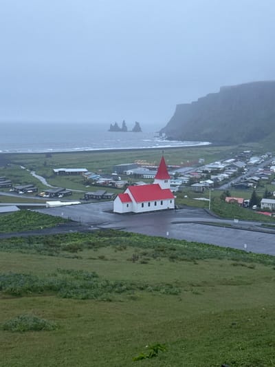 The image depicts the town of Vik, Iceland, on a possibly overcast day. A distinctive white church with a red roof is prominently featured in the foreground, alongside a paved road. The town itself, consisting of various buildings and houses, stretches towards a dark, rocky coastline with sea stacks visible in the distance. To the right of the church is a large green hillside or field. Fog or mist is apparent in the distance obscuring the cliffs.