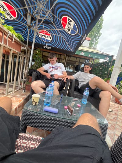 The image depicts three young men relaxing at an outdoor cafe or restaurant patio in Slashten, Bulgaria, during what appears to be a bright daylight hour, possibly overcast. The scene is viewed from an elevated, oblique perspective.
In the foreground and to the right, the legs of an individual wearing black shorts are visible, resting on a checkered cushion. A large, black and blue umbrella, featuring the "PEPSI" logo repeatedly, provides shade over the left side of the frame.
Two other young men are seated around a dark wicker glass-top table. One man, wearing a light grey t-shirt and dark shorts, is leaning back in his chair with his legs crossed, looking towards the camera. The other, dressed in a white graphic t-shirt and black shorts, is focused on his phone, holding it with both hands.
The glass table holds several items: two plastic bottles of "DEVIN" branded water, a brown box labeled "TERREA", a purple rectangular case, a yellow paper cup with a design, and some small pink and white plastic items.
In the background, a green banner is visible with Bulgarian text: "КАМЕНИЦА" (Kamenitza, a Bulgarian beer brand) and "МЪЖЕТЕ ЗНАЯТ ЗАЩО" (Men know why). Beyond the patio structure, green foliage and trees are visible against a bright sky. The flooring of the patio consists of light brown and reddish-brown tiles. The overall impression is one of casual leisure.